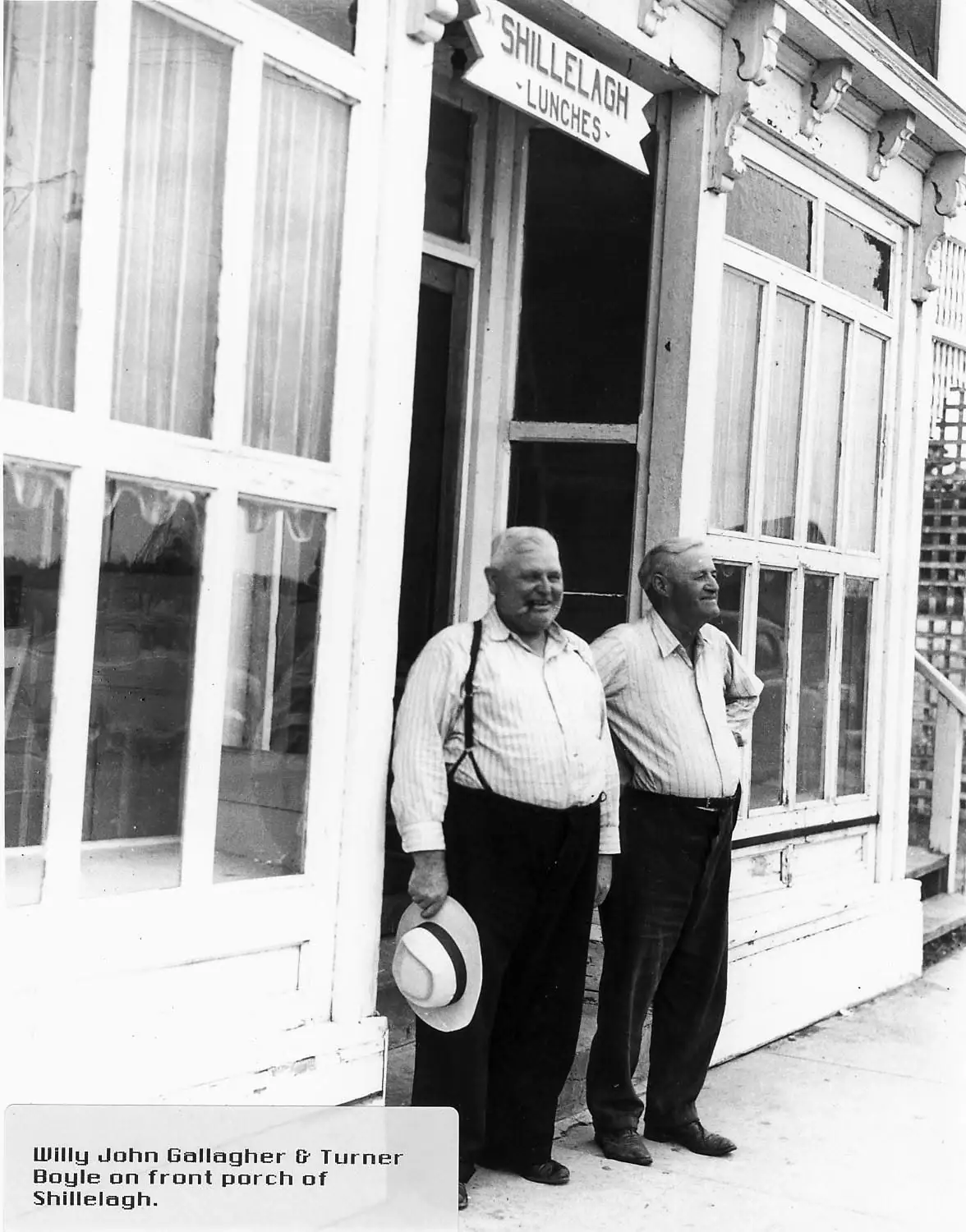 Two men stand on the porch of a storefront beneath a sign reading “SHILLELAGH - LUNCHES -.”