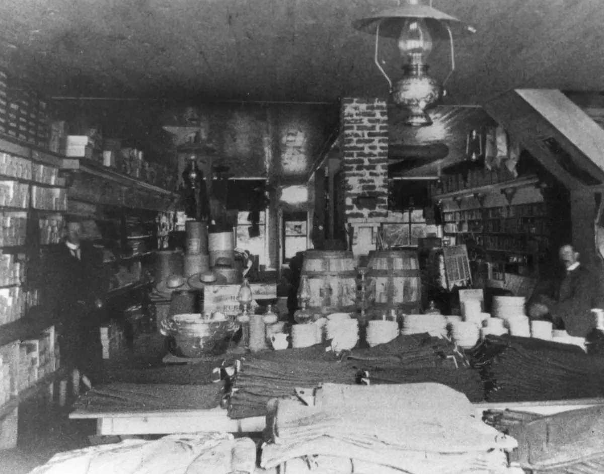 Interior of a crowded store with shelves, barrels, merchandise, a hanging lamp, and two men.