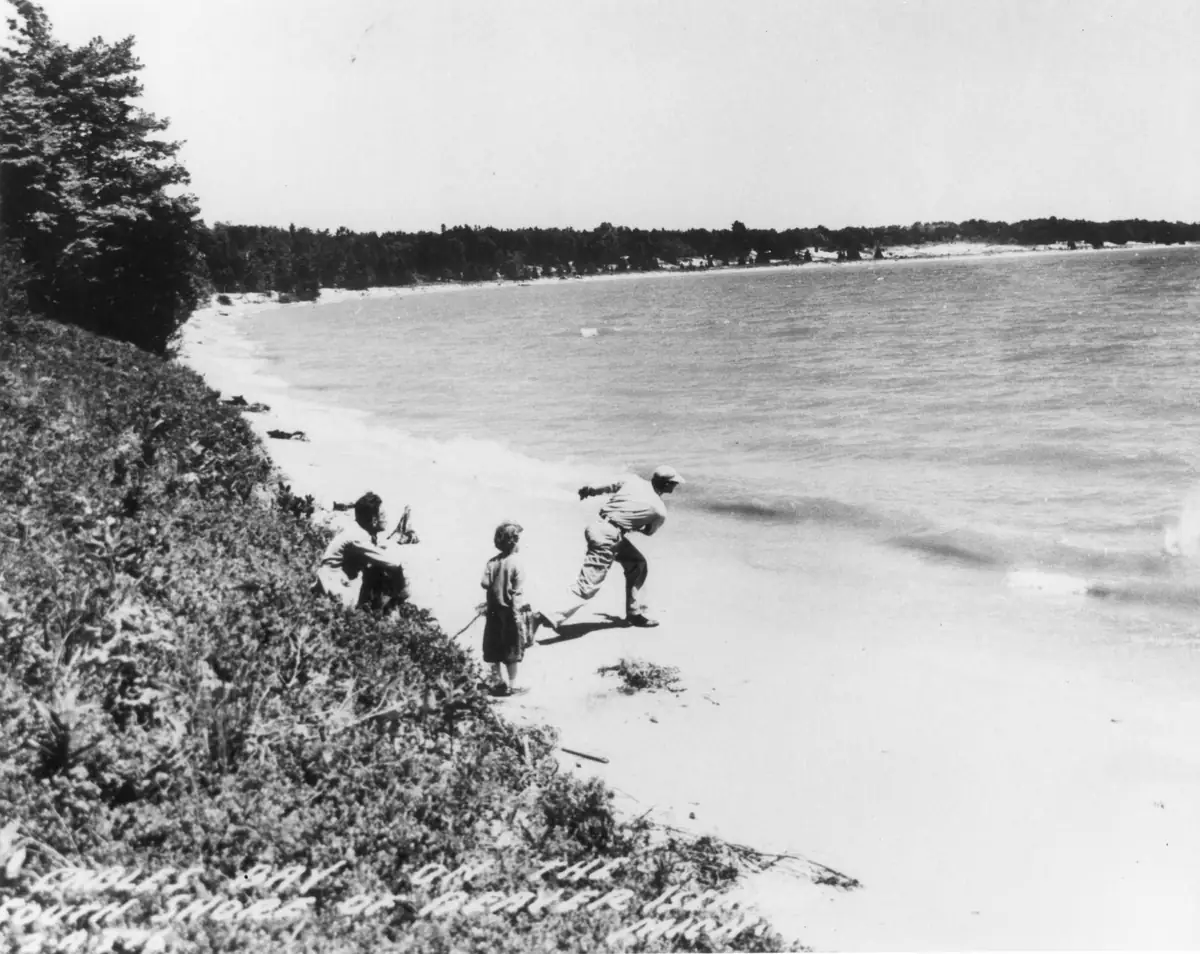 Three people stand and sit on a sandy shoreline as one appears to skip a stone across the water.