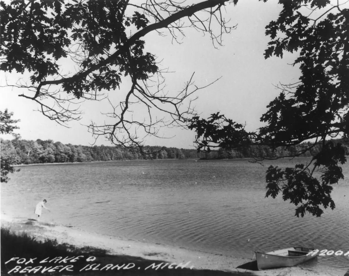 Black-and-white shoreline photograph with a wading person, canoe, wooded far shore, and handwritten caption.