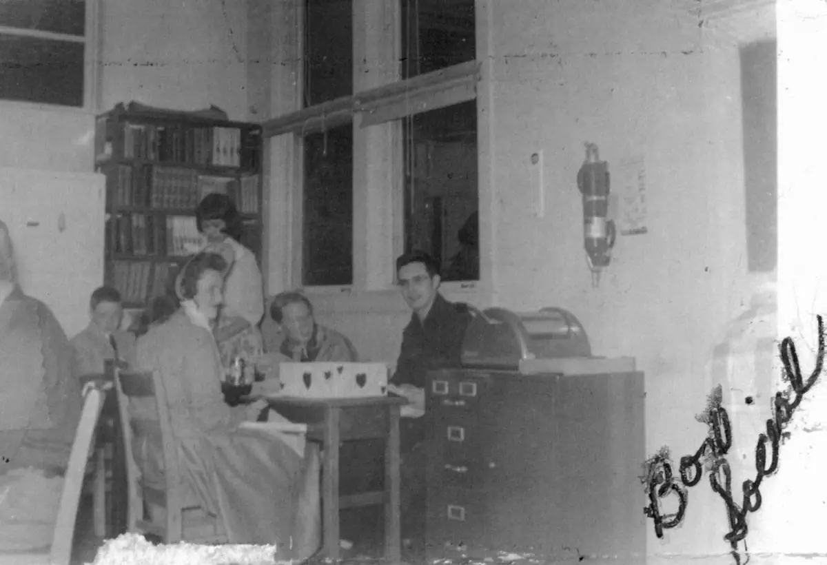 Black-and-white photograph of several people gathered around a table in a room with bookshelves and filing cabinets.