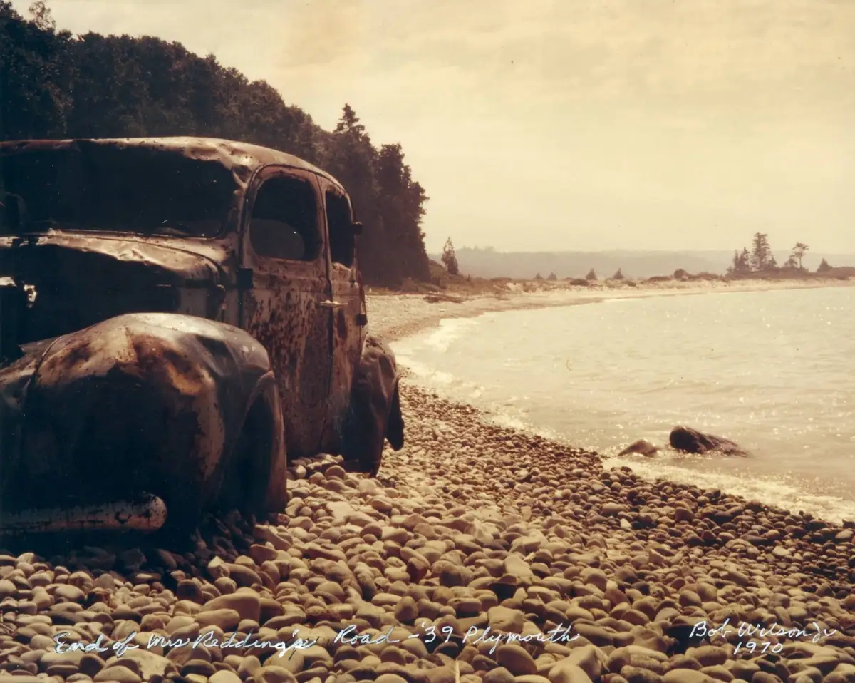 A heavily rusted old car sits on a rounded-stone shoreline beside calm water and trees.