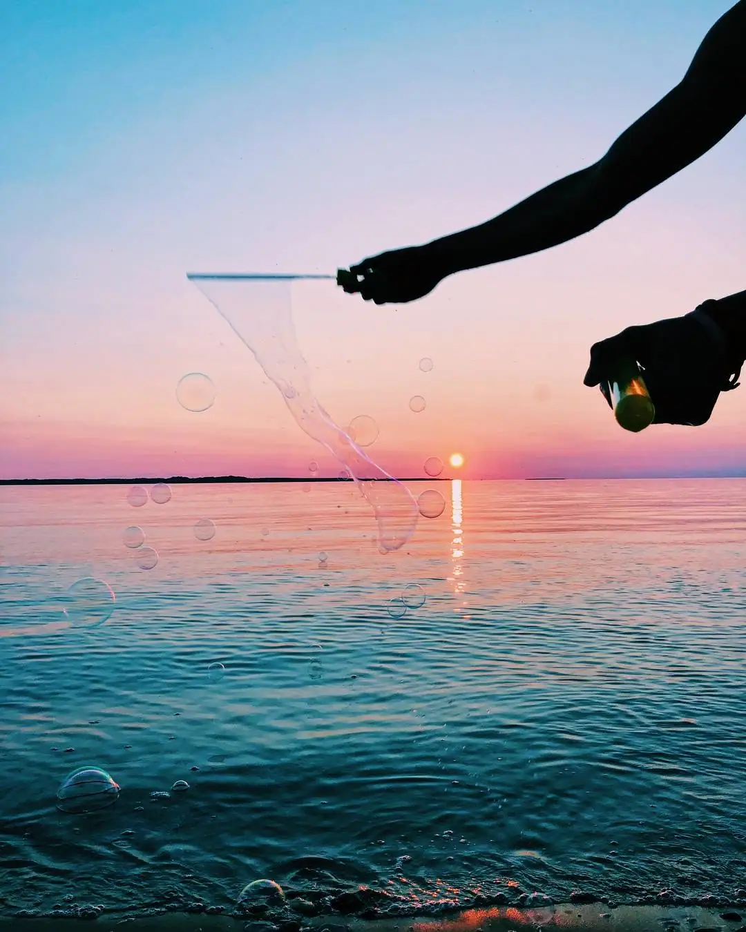 Silhouetted hands make bubbles over calm water at sunset.