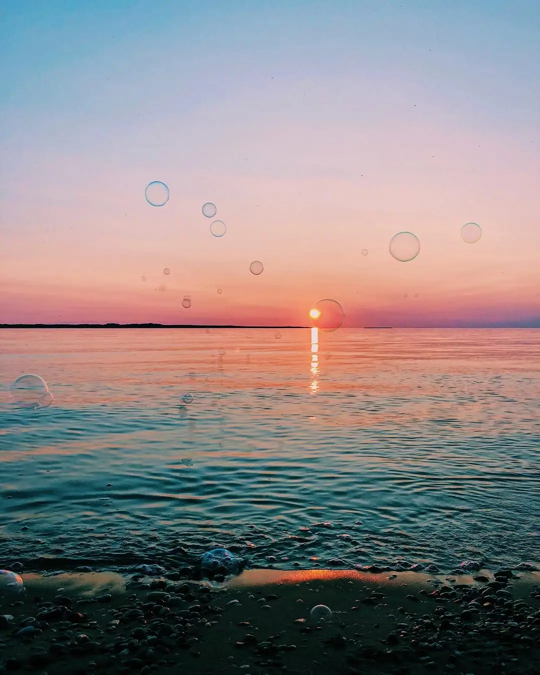 Sunset over rippling water with bubbles above a pebbled shoreline.
