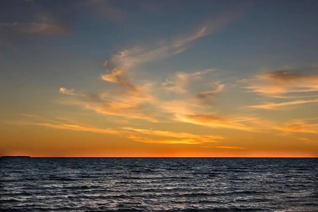 Sunset colors the horizon above dark, rippling open water beneath streaks of wispy clouds.