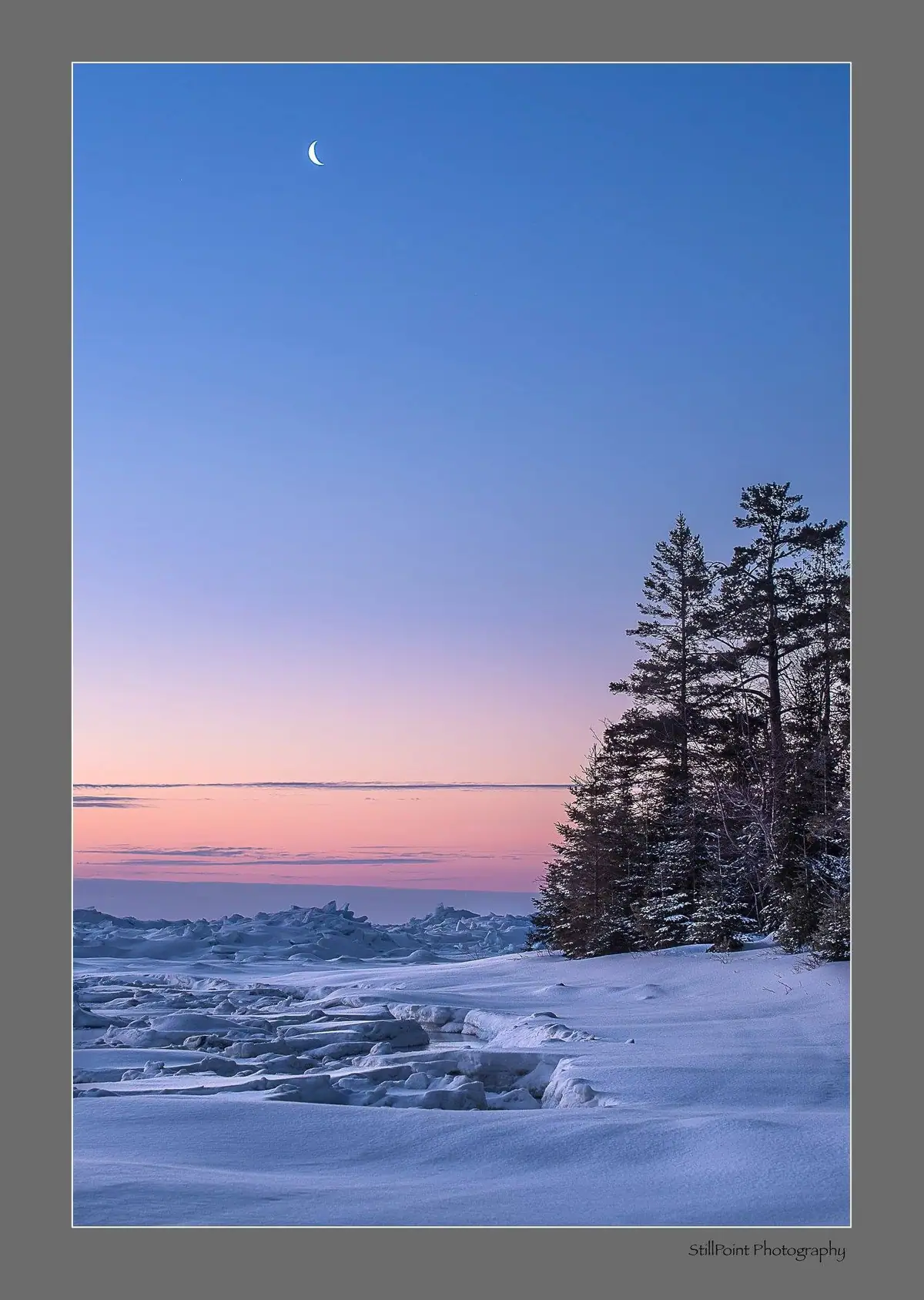 Snow-covered shoreline and ice formations beneath a crescent moon and pastel dawn sky, with evergreen trees at right.
