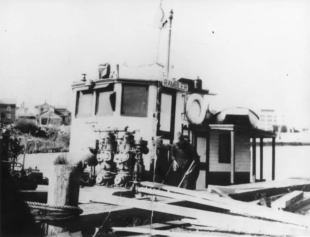 Black-and-white photograph of a small workboat with exposed machinery beside a wooden pier.