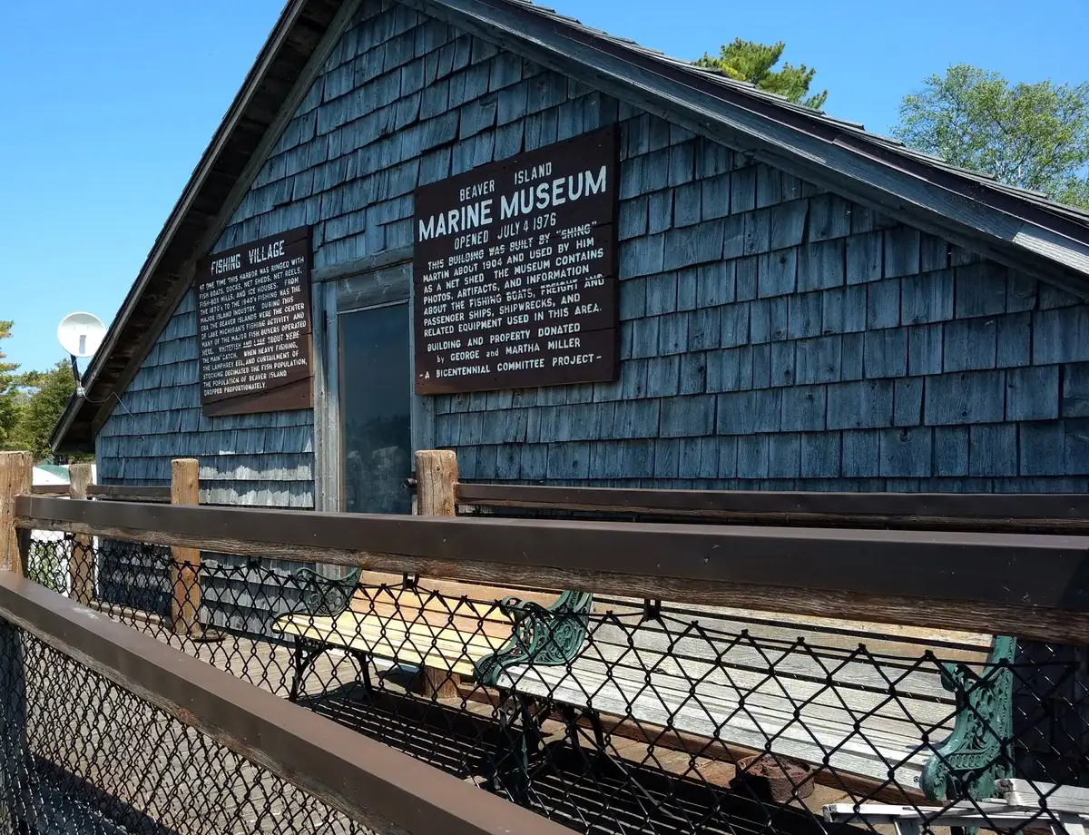 A shingle-sided wooden building with two historical signs, viewed behind a chain-link fence and wooden railing.