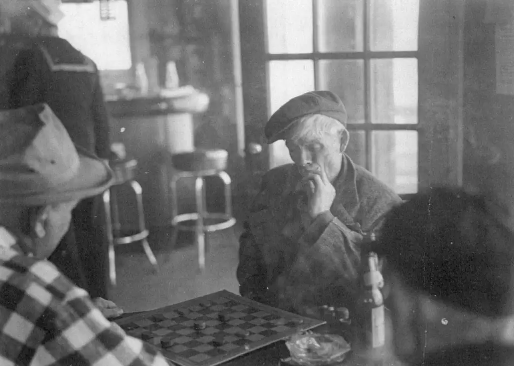 Two men sit across a table, playing a board game in a room with a large multi-pane window.