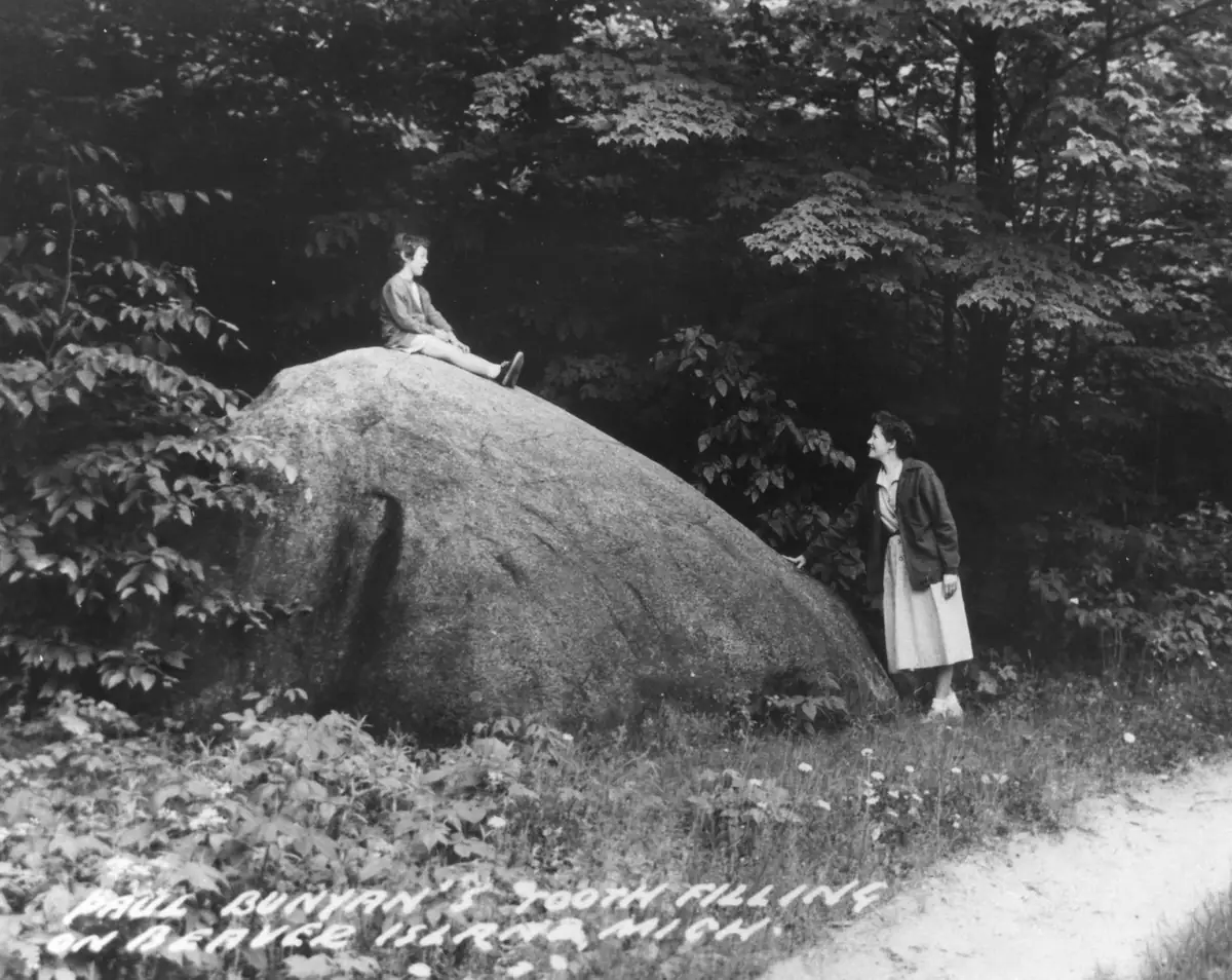 A child sits atop a large boulder while a woman stands beside it in a wooded setting.