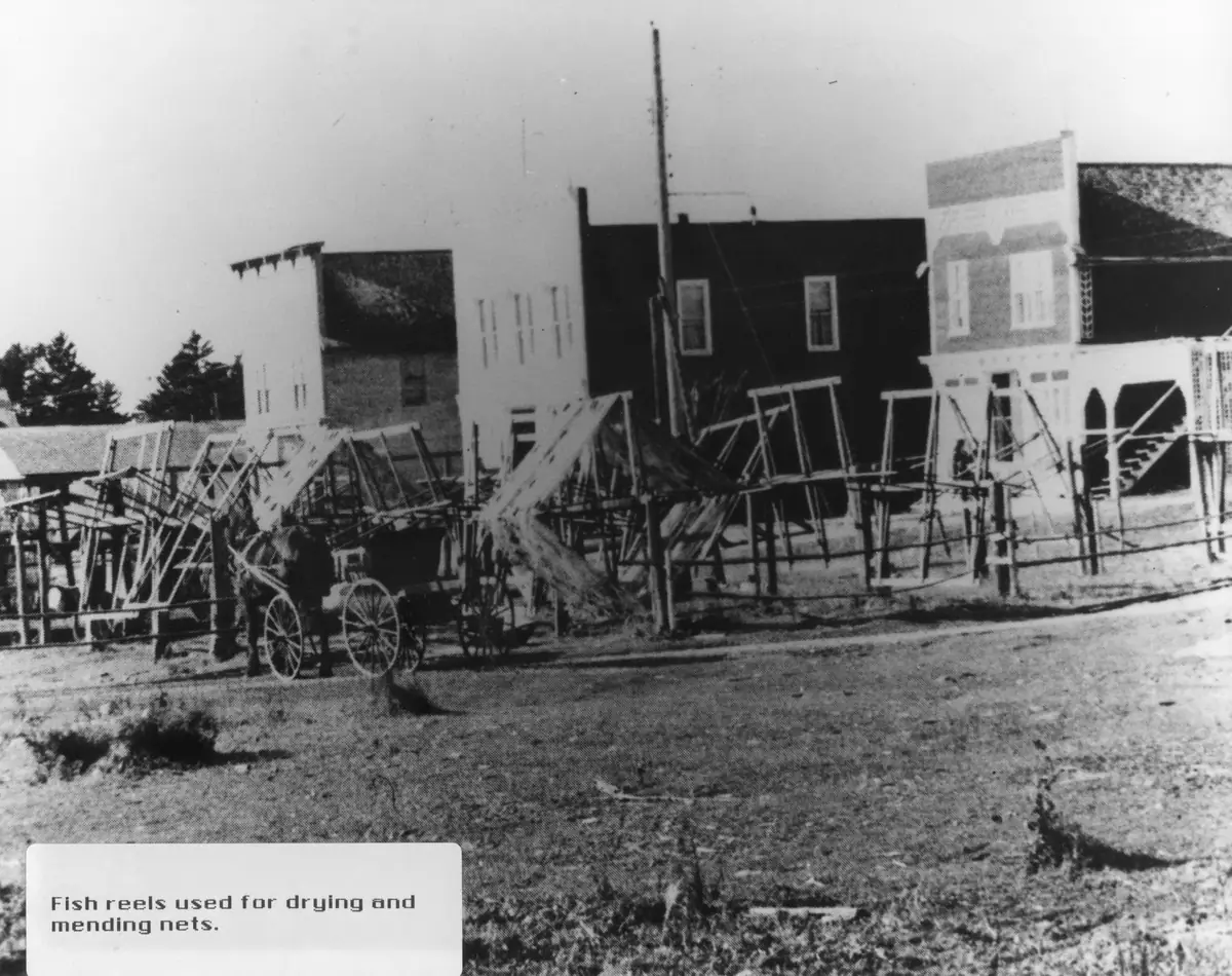 Fish reels and a horse-drawn cart line a dirt street beside early commercial buildings.