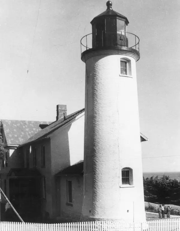 Black-and-white photograph of a cylindrical lighthouse beside a roofed building and picket fence.
