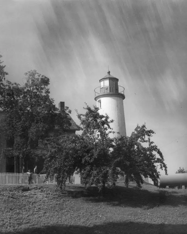 Black-and-white photograph of a cylindrical lighthouse beside a tree-shaded house and picket fence.