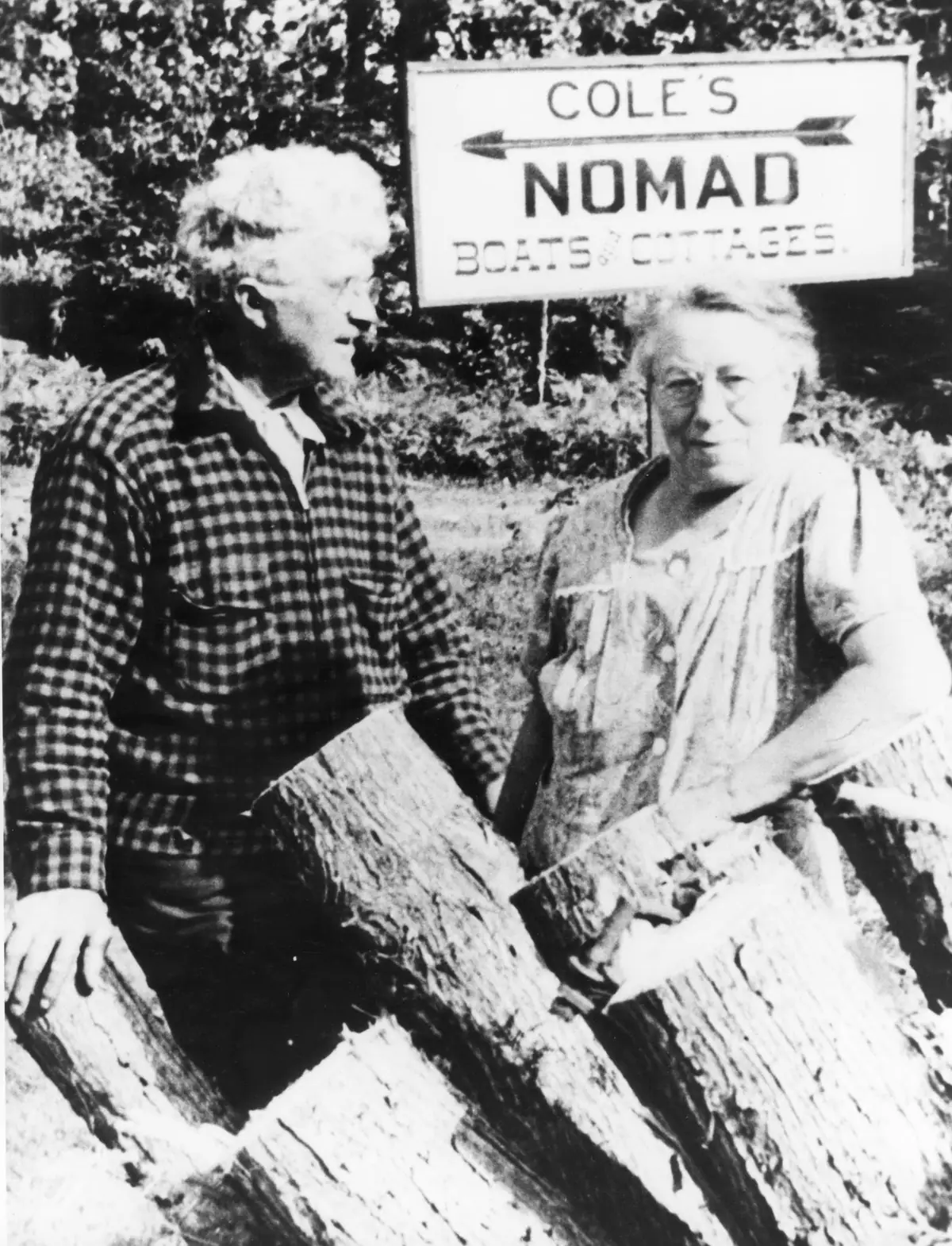 Two adults pose beside cut logs beneath a sign reading “COLE'S NOMAD BOATS COTTAGES.”