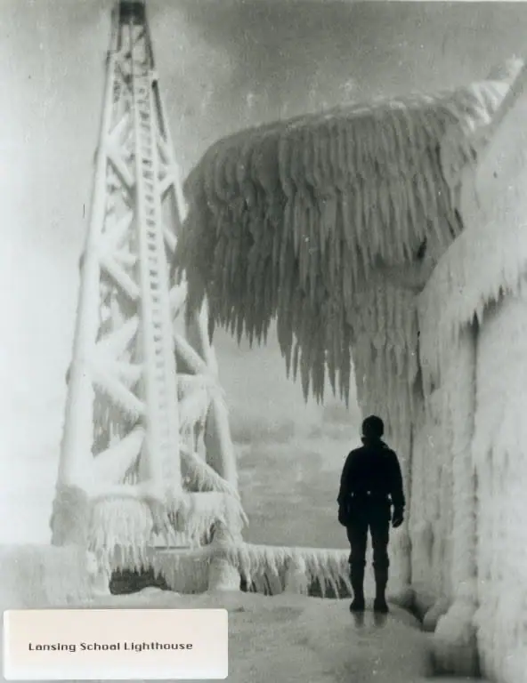 Black-and-white photograph of an ice-covered lighthouse tower with a person standing beside it.