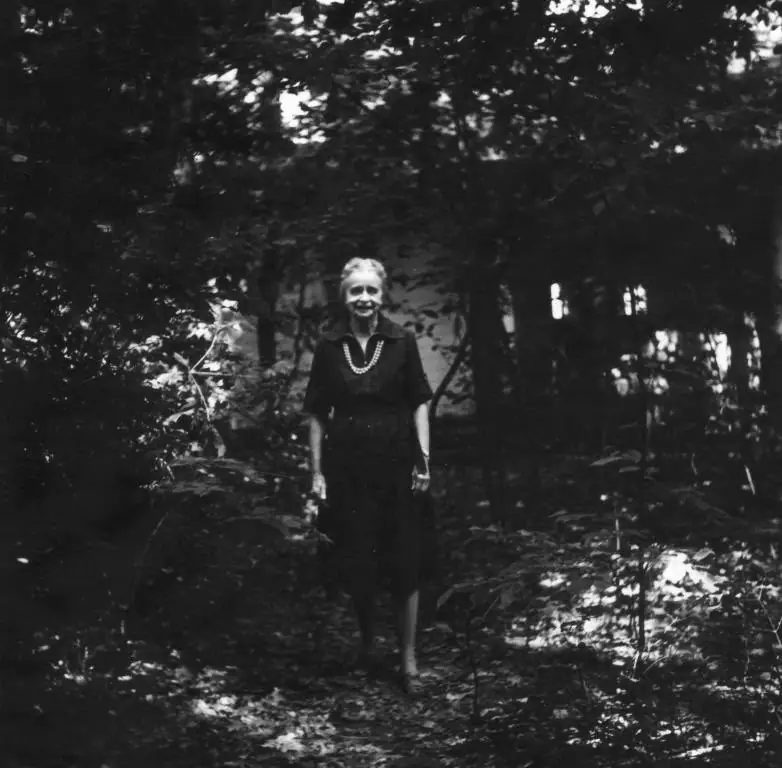 Black-and-white photograph of an older woman standing on a shaded path among trees.