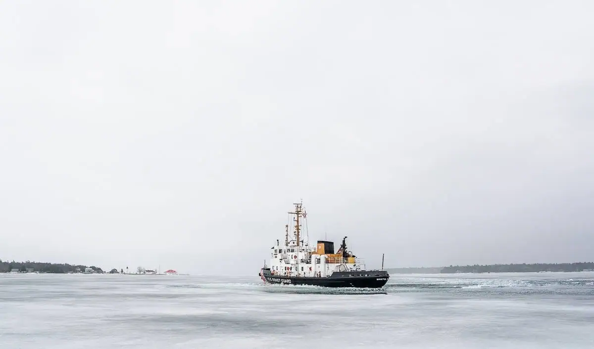 A black-and-white cutter moves through broken ice beneath an overcast sky, with a distant wooded shoreline behind it.