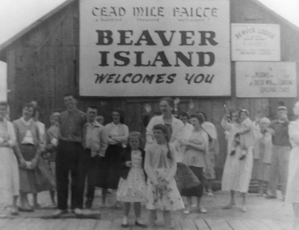 A group of adults and children poses beneath a large welcome sign outside a wooden building.