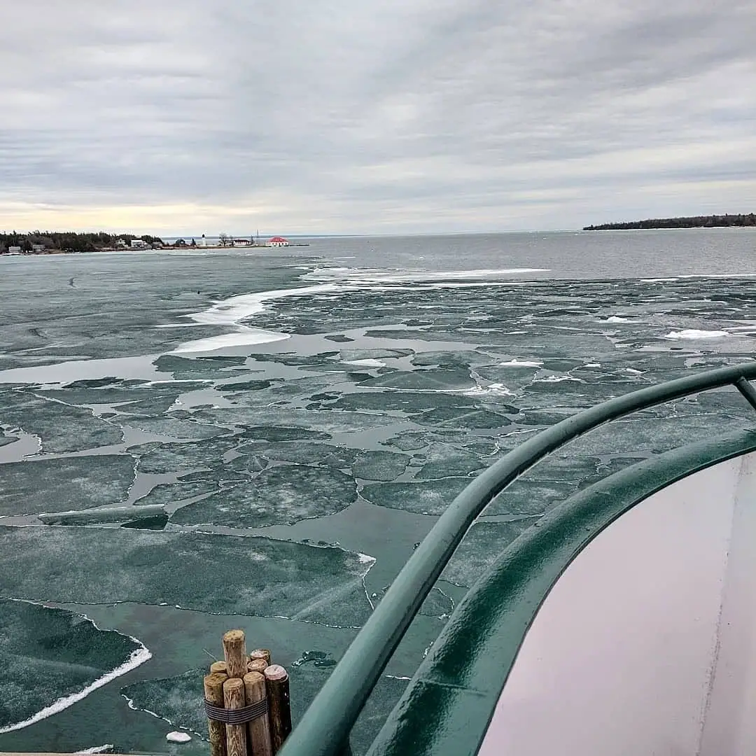 A vessel moves through broken ice toward a distant shoreline with a lighthouse and waterfront buildings.