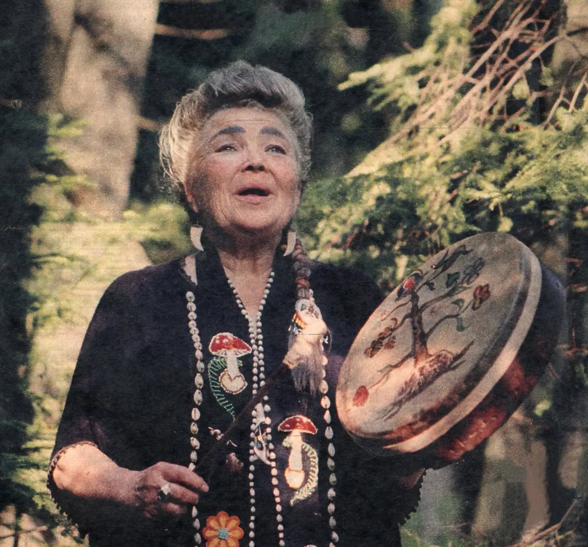 Portrait of an older woman holding a large decorated hand drum outdoors among trees.