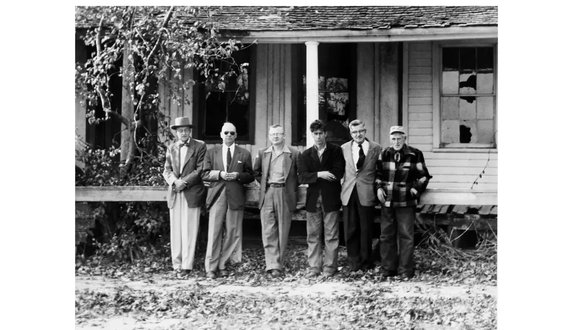 Six men pose in front of a weathered wooden house with a covered porch in a black-and-white photograph.