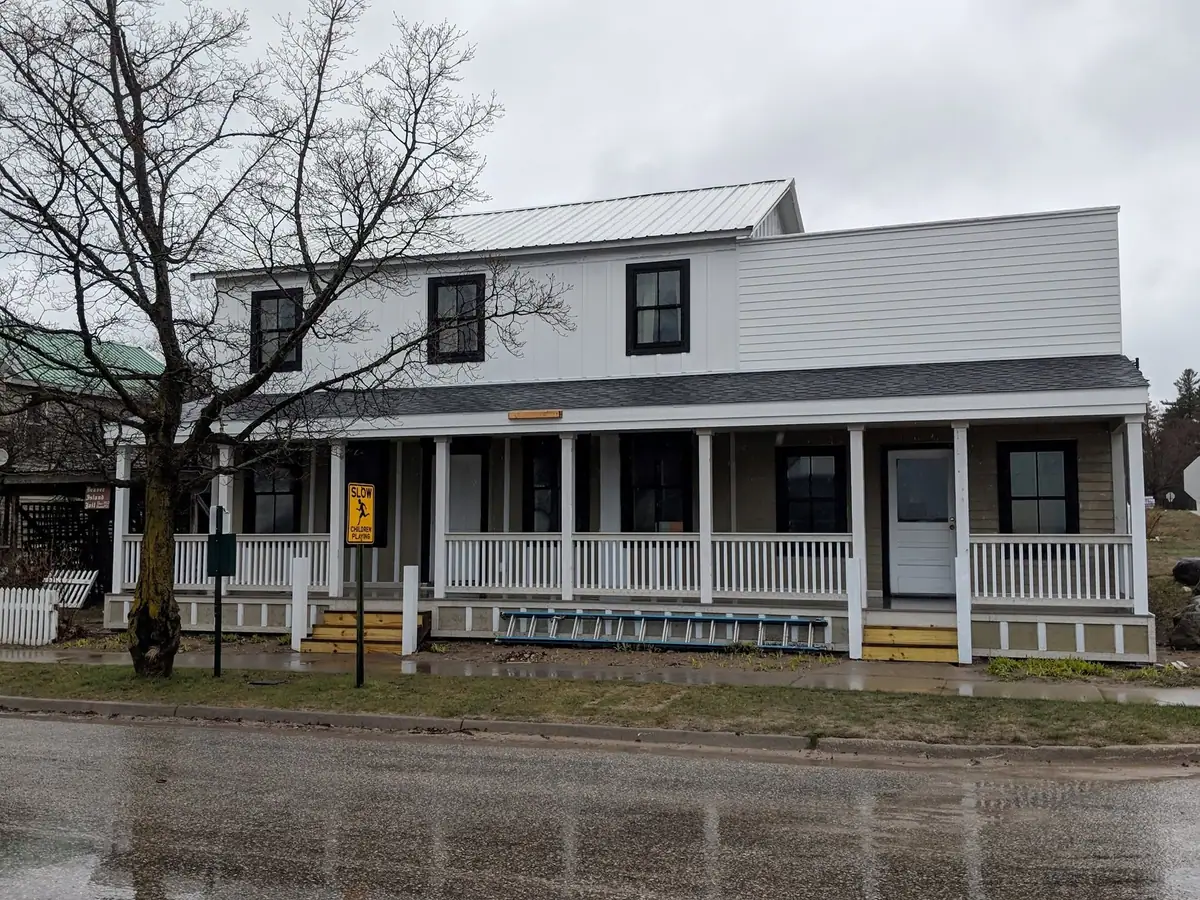 A white two-story building with a covered porch, roadside warning sign, ladder, and leafless tree.