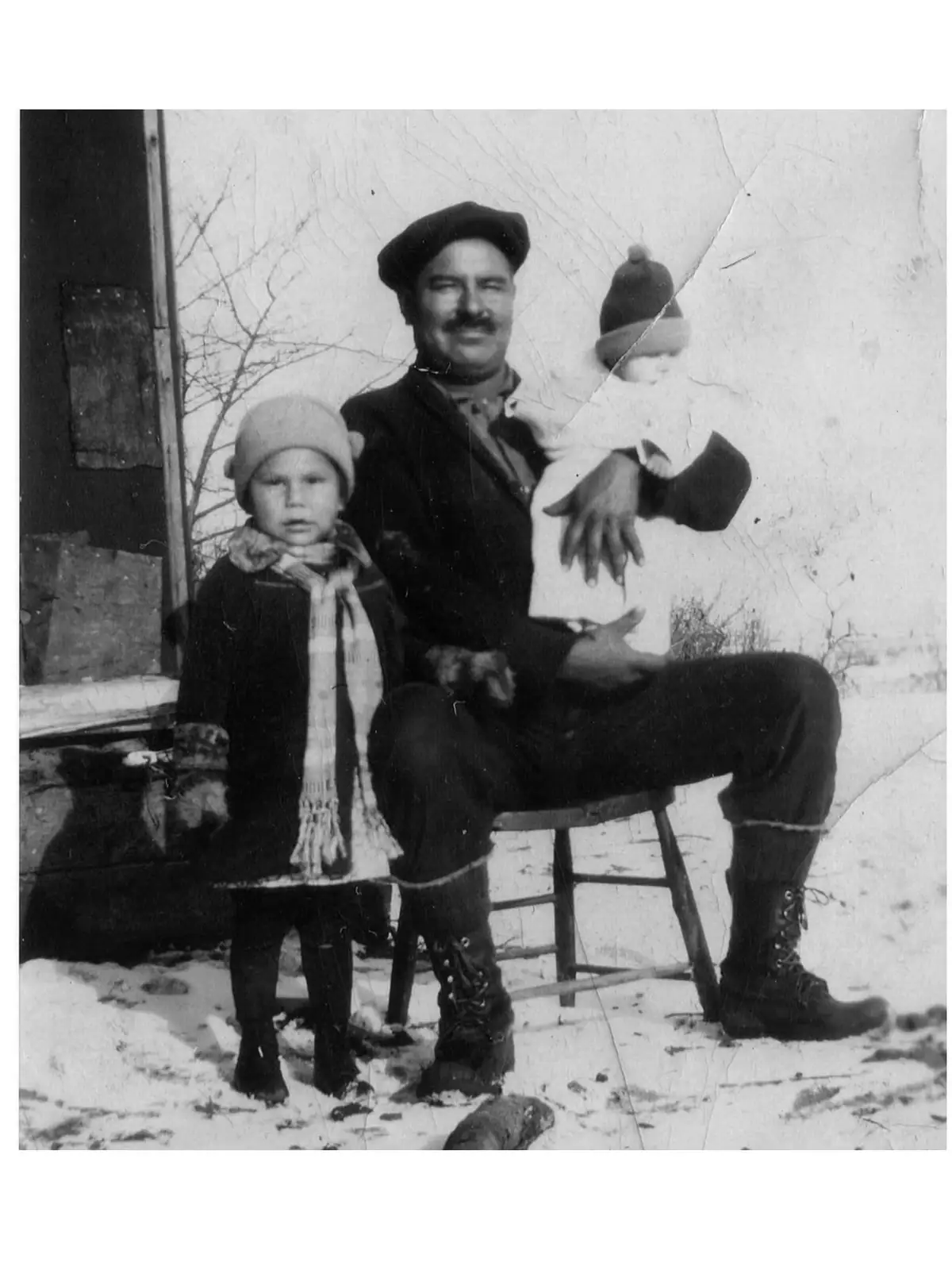 A man sits outdoors in the snow holding a bundled infant, beside a bundled child in winter clothing.