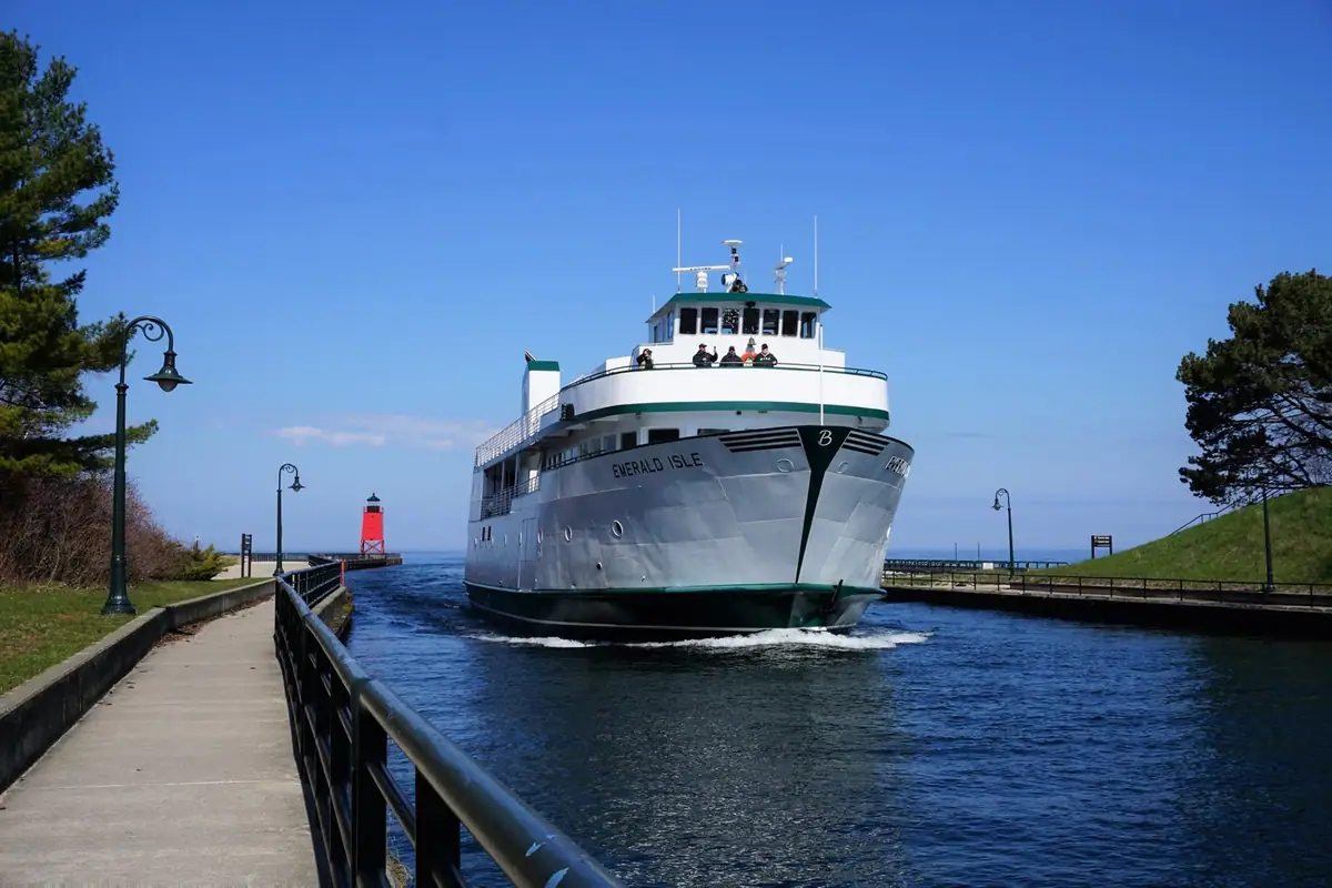 A passenger ferry moves through a canal beside walkways, railings, trees, and a red lighthouse.