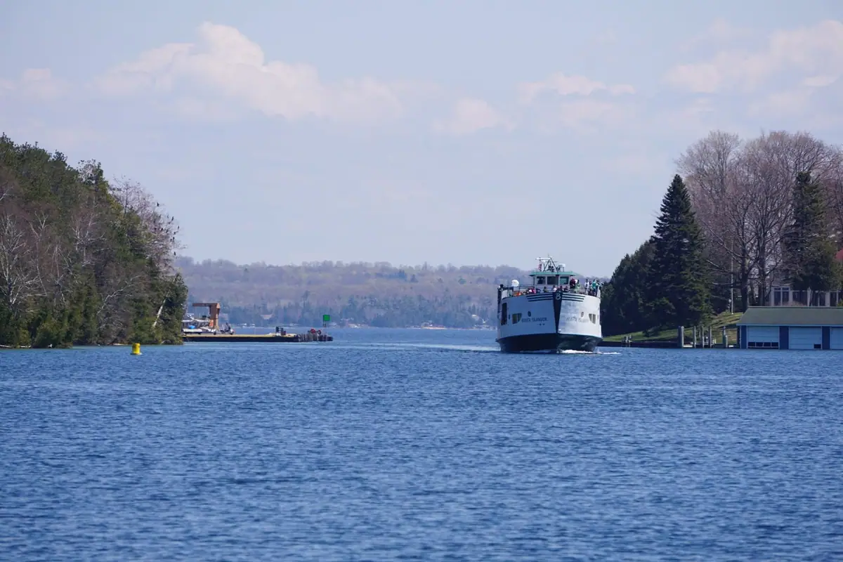 A white ferry approaches along a tree-lined waterway near docks and shoreline buildings.