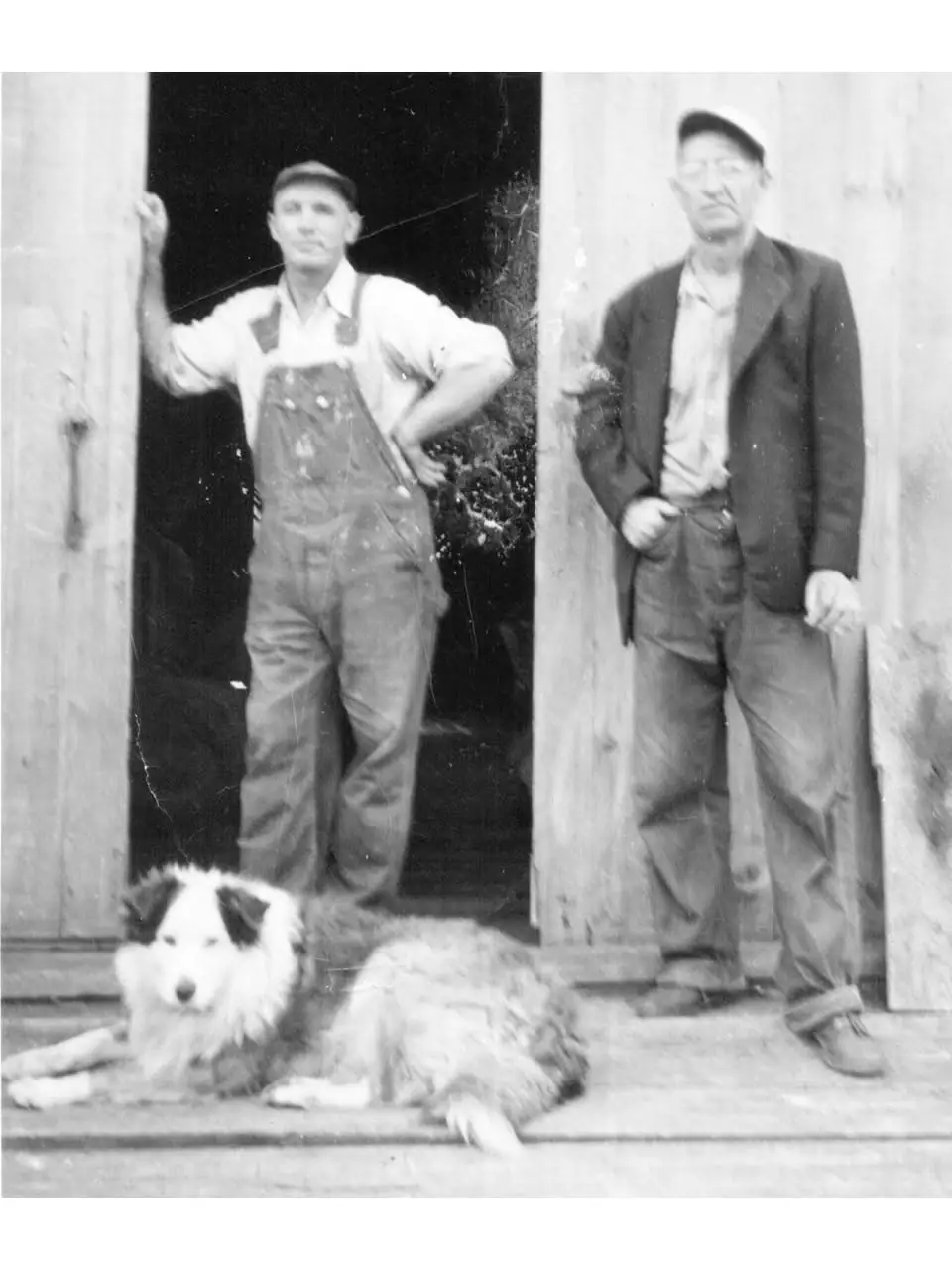 Two men and a dog pose in the doorway of a wooden building in a black-and-white photograph.