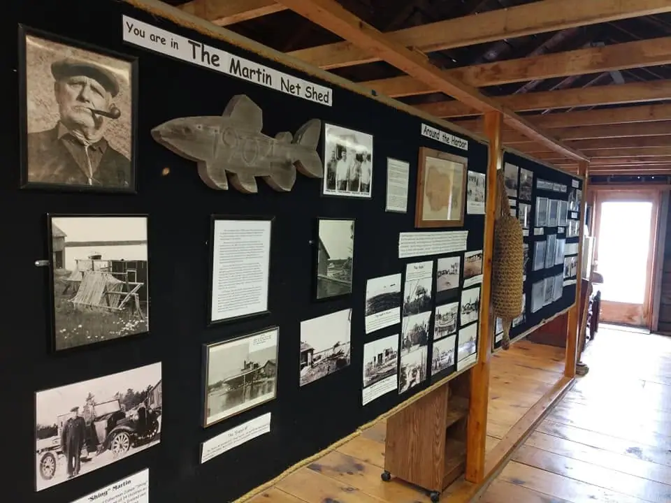 Interior of a wooden exhibit space with framed photographs, text panels, a carved fish, and a hanging net.