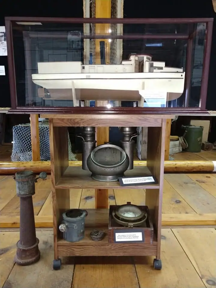 A model boat and maritime equipment are displayed in a wooden and glass museum case.