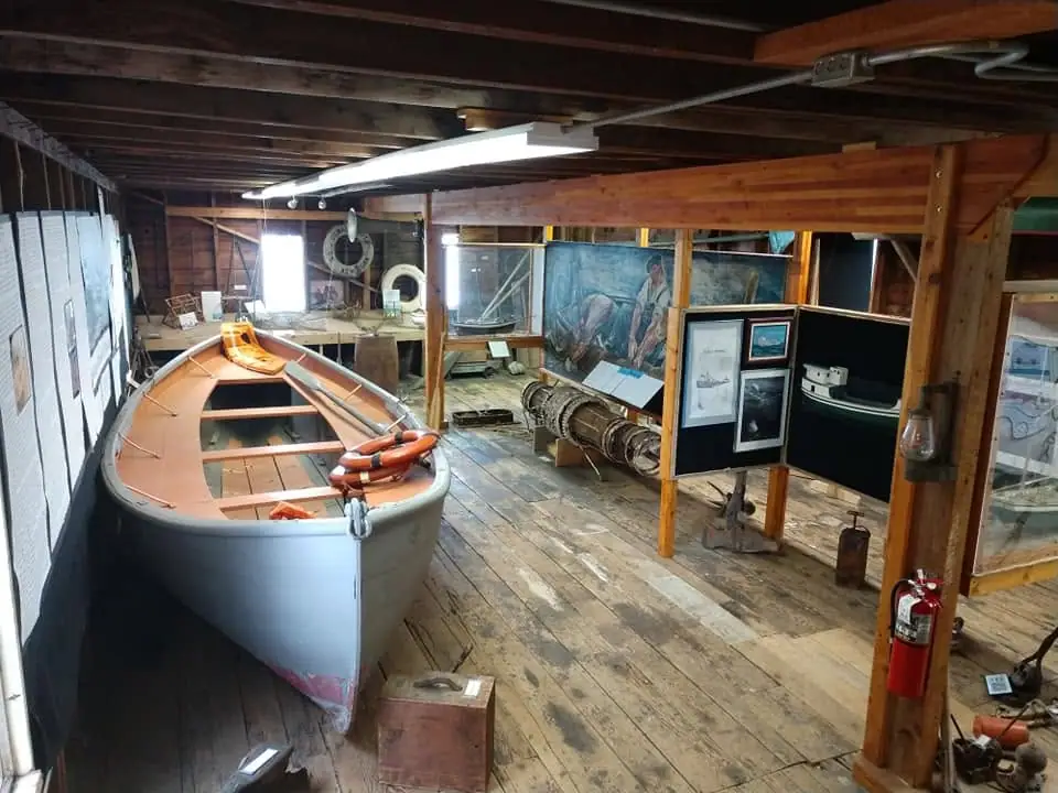 Interior exhibit space with a small boat, maritime objects, framed displays, and a large painting.