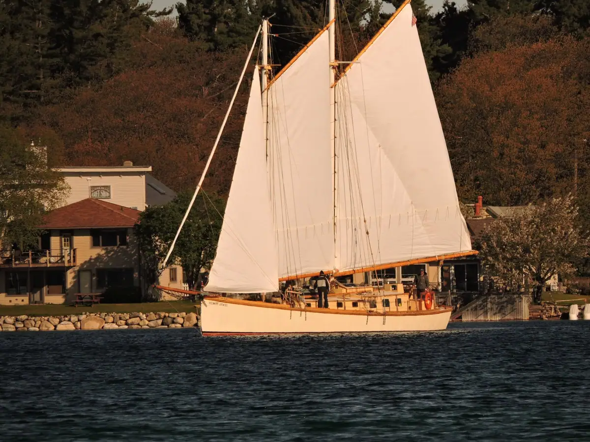 Two-masted schooner under full sail beside a wooded shoreline and waterfront buildings.