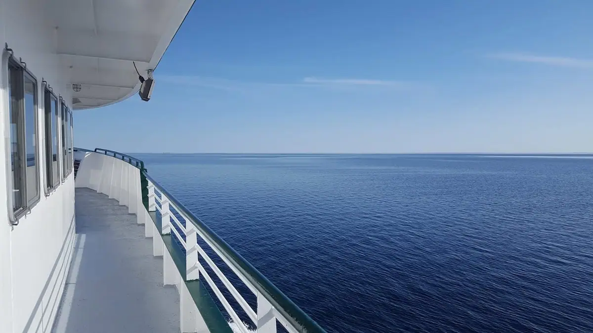 View along a passenger vessel’s side deck toward calm open water beneath a clear blue sky.