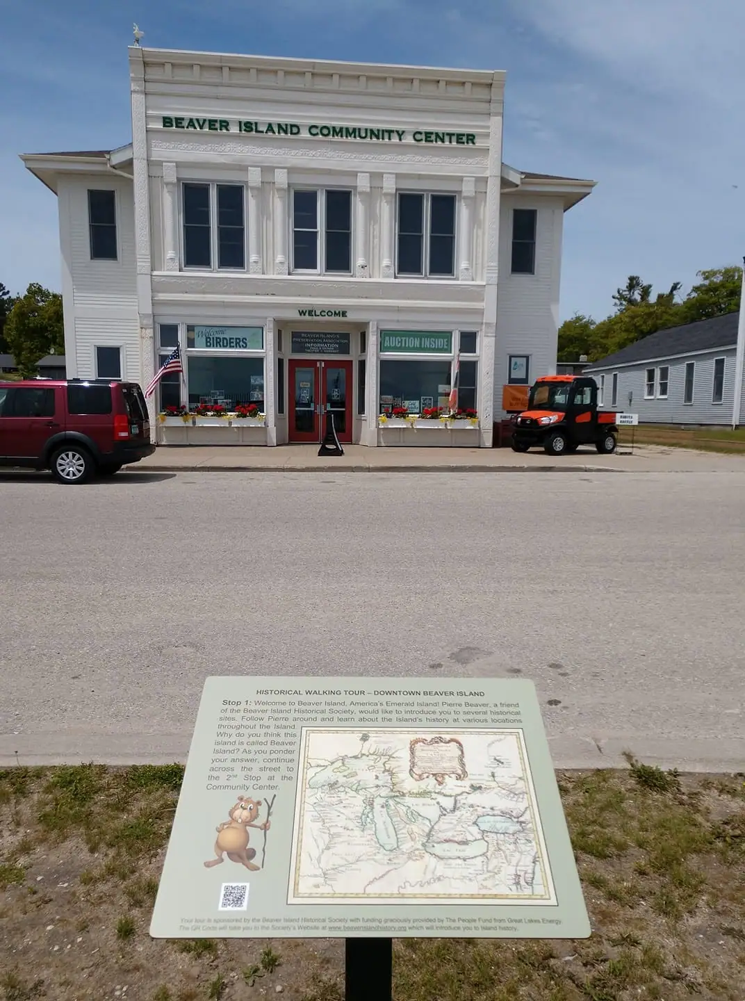 A white community center with storefront signs, parked vehicles, and a historical walking-tour panel in front.