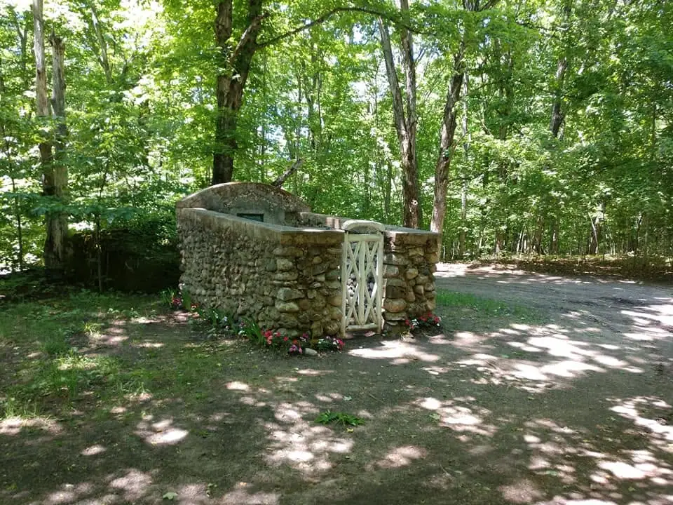A low stone enclosure with a white gate stands among trees beside a gravel path.