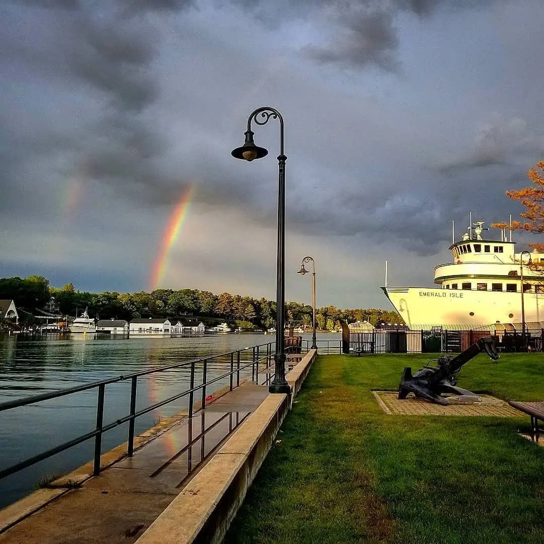 A white ferry, waterfront walkway, and two rainbows beneath dark clouds.