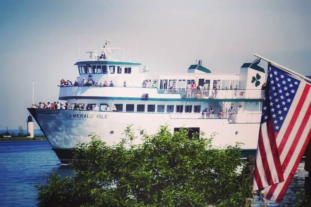 A white passenger ferry crowded with people passes behind leafy shrubs beside a large American flag.