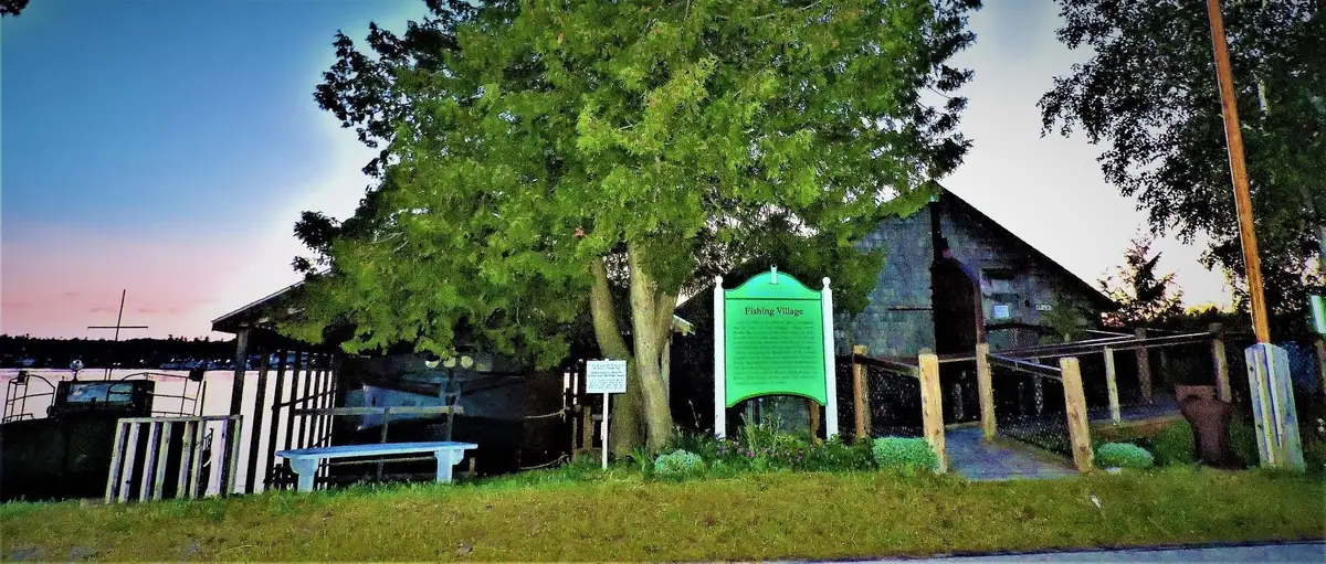 A green historical marker stands beside a large tree and weathered wooden waterfront buildings at dusk.