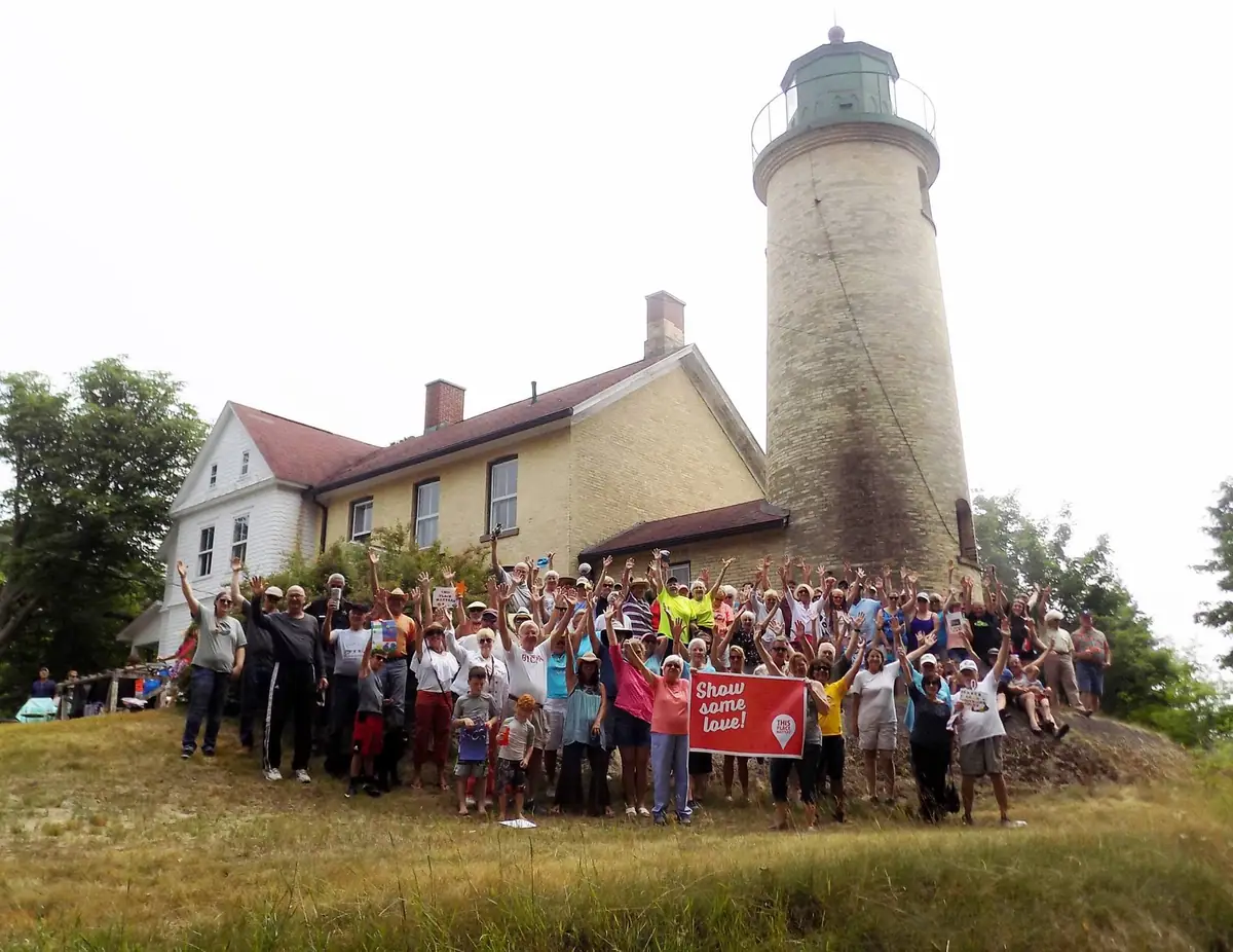 A large group poses with raised arms in front of a lighthouse and keeper’s buildings, holding a red banner.