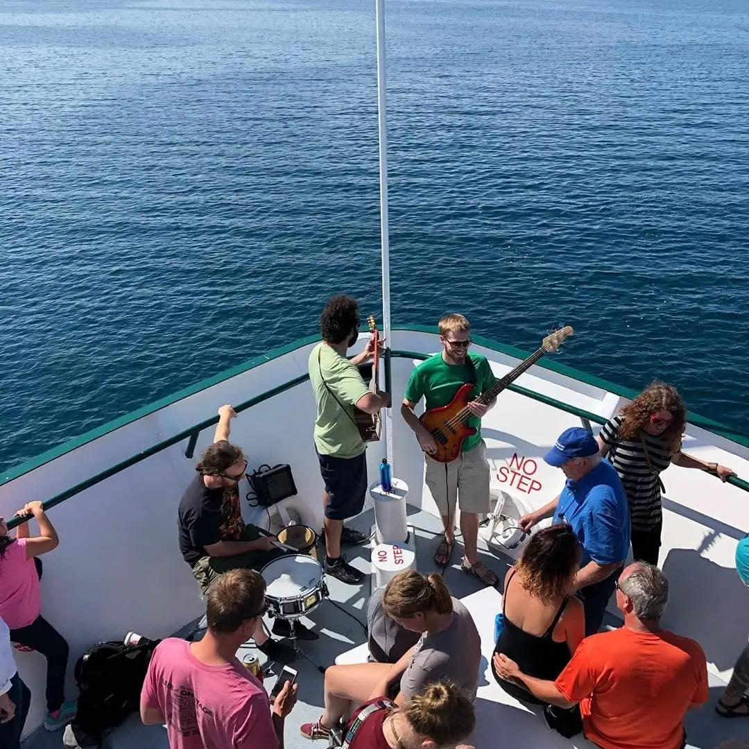 Musicians perform on a passenger boat’s open deck while people gather around them.