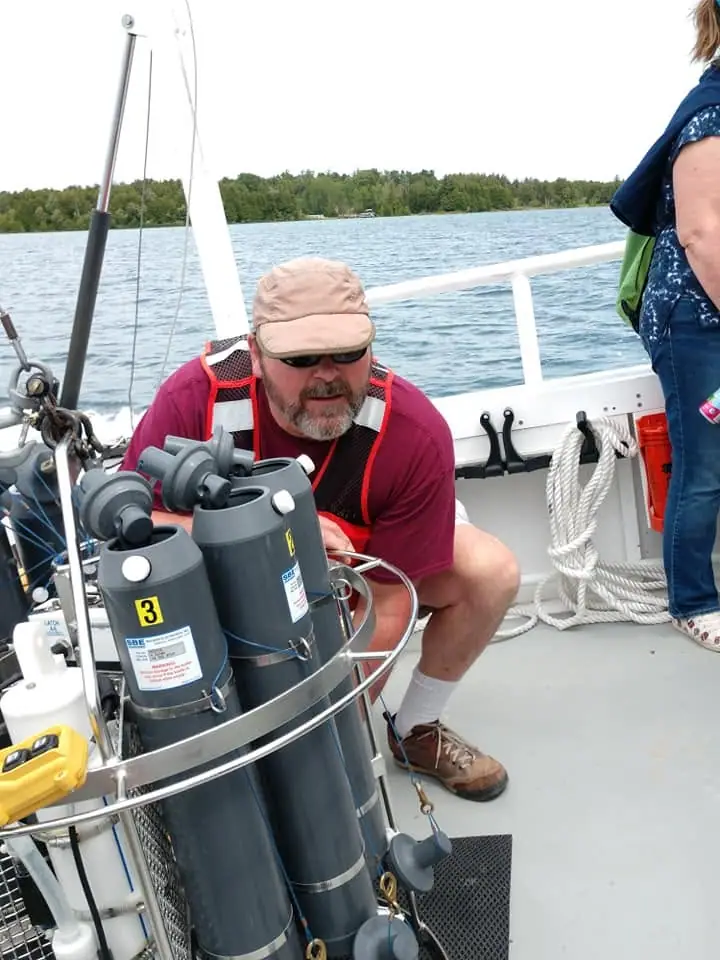 A person in a red life vest crouches beside cylindrical water-sampling equipment on a boat deck.