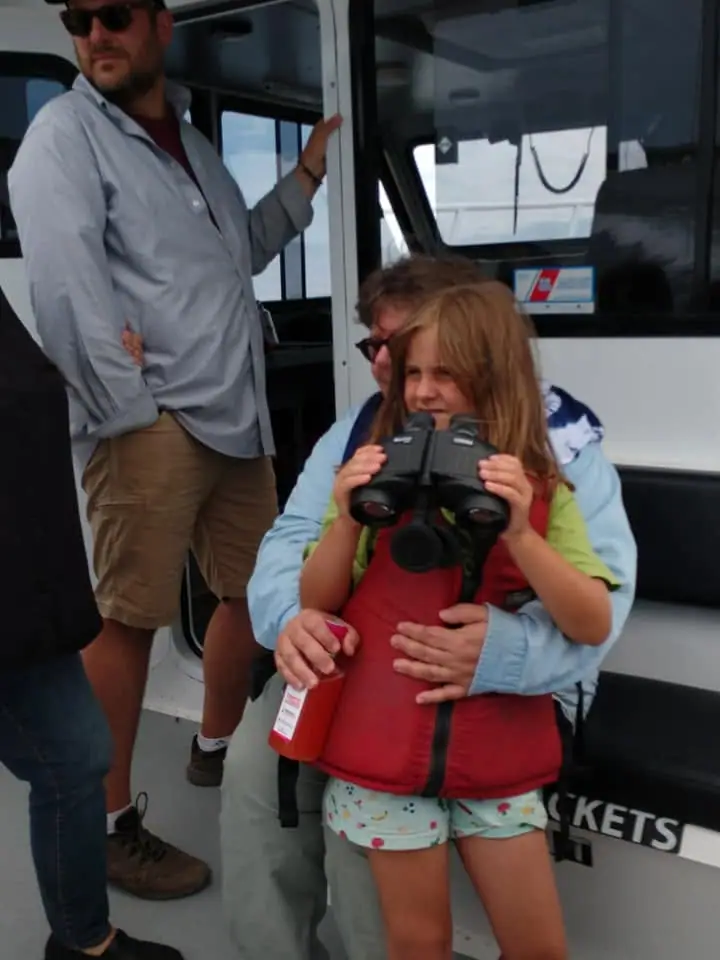 A child in a red life vest looks through binoculars while an adult holds her inside a boat.