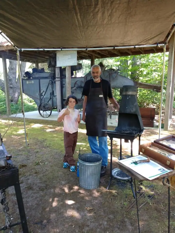 An adult and child stand beneath a canopy beside historic machinery and a metal furnace.