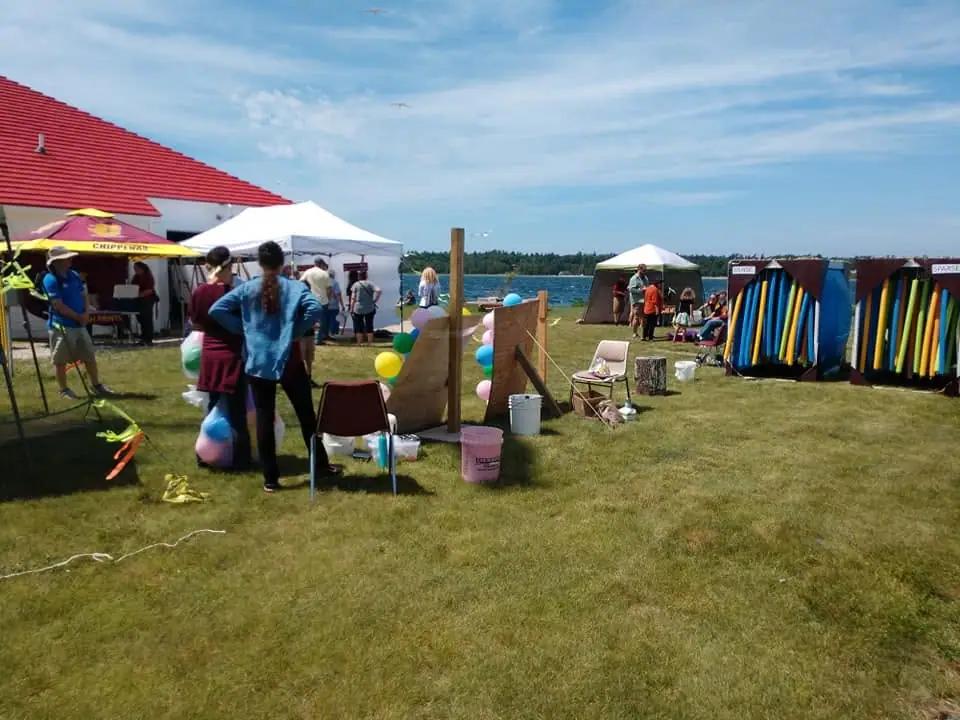 People gather at a grassy waterfront event with tents, food service, balloons, and colorful game stalls.