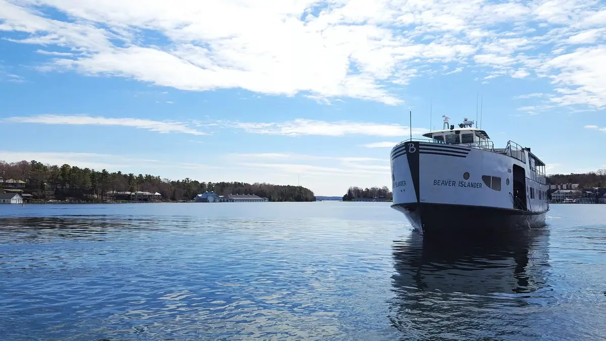 A white ferry approaches across calm water beneath a bright, cloud-streaked sky.