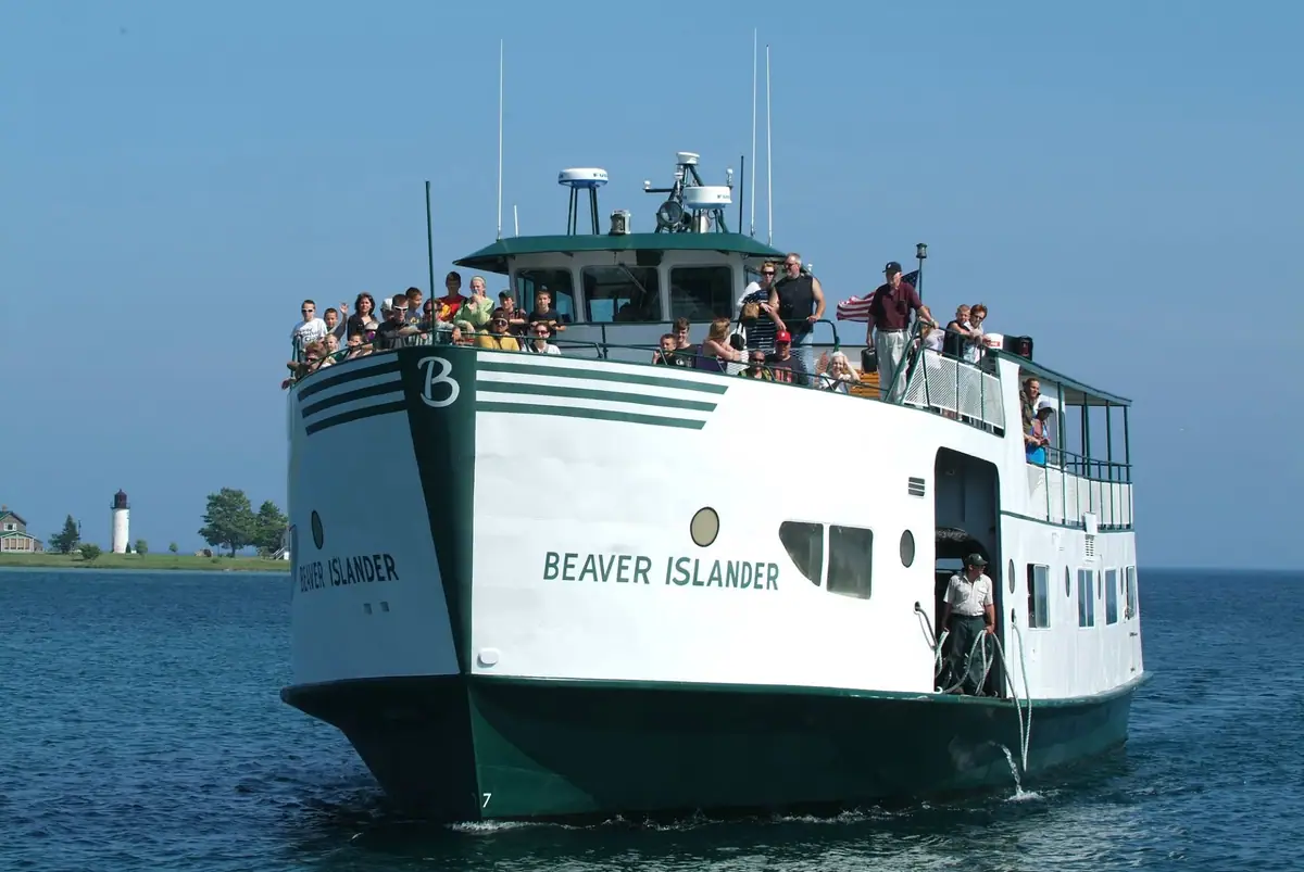 A crowded white-and-green passenger ferry approaches on open water with a lighthouse visible behind it.
