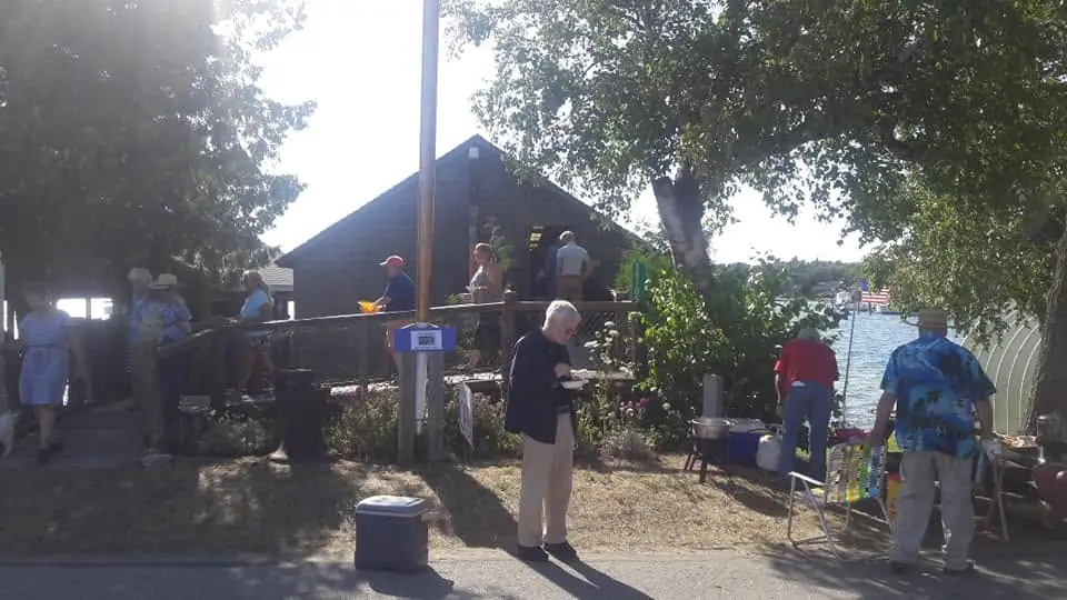 People gather beside a dark wooden cabin and waterfront, with folding chairs, coolers, and a small boat visible.