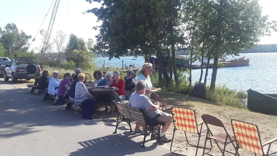 People gather outdoors at a picnic table beside the water, with a boat, crane, vehicles, and trees behind them.